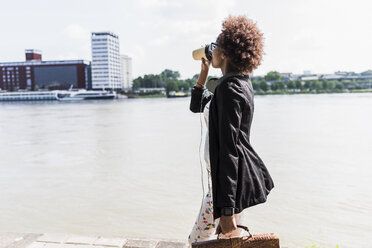 Germany, Mannheim, young businesswoman drinking coffee to go while walking at riverside - UUF007769