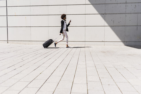 Walking businesswoman with trolley bag looking at her smartphone - UUF007774