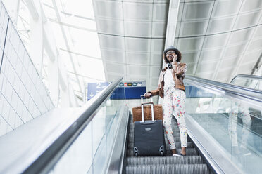 Young woman with baggage standing on escalator telephoning with smartphone - UUF007775