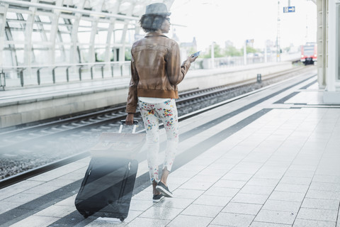 Rückenansicht einer jungen Frau mit Smartphone und Gepäck am Bahnsteig, lizenzfreies Stockfoto