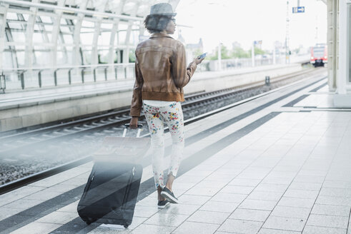 Rückenansicht einer jungen Frau mit Smartphone und Gepäck am Bahnsteig - UUF007777