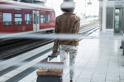 Rückenansicht einer jungen Frau mit Rollkoffer und Aktentasche am Bahnsteig - UUF007779