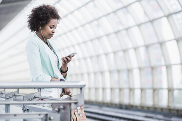 Junge Frau mit Kopfhörern schaut auf ihr Smartphone, während sie am Bahnsteig wartet - UUF007785