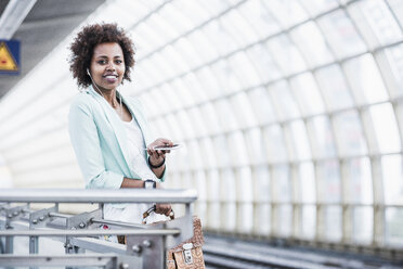 Portrait of young woman with earphones and smartphone waiting at platform - UUF007786