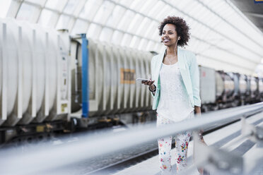 Portrait of smiling young woman with earphones and smartphone at platform - UUF007791