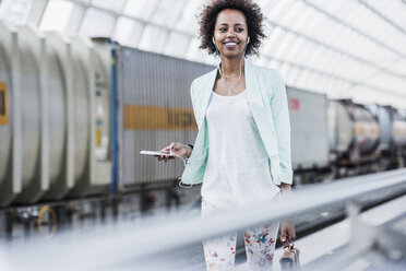 Portrait of smiling young woman with earphones and smartphone at platform - UUF007792