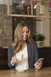 Portrait of smiling young woman in a coffee shop looking at her smartphone - KAF000158