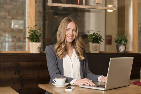 Portrait of smiling young woman using laptop in a coffee shop - KAF000159