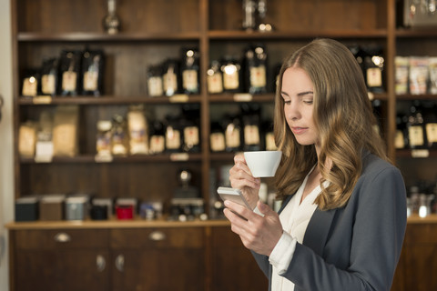 Junge Frau trinkt Kaffee in einem Café und schaut auf ihr Smartphone, lizenzfreies Stockfoto