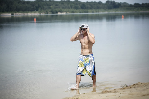 Mann in Badehose hört Musik mit Kopfhörern beim Waten am Seeufer, lizenzfreies Stockfoto