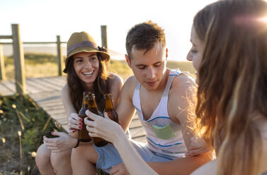 Friends sitting on boardwalk at the beach, drinking beer at sunset - MGOF001986