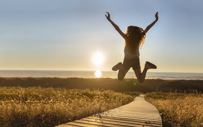 Young woman jumping on boardwalk at the beach with raised arms - MGOF001991