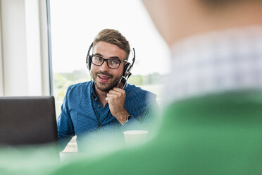 Mann mit Headset und Laptop im Büro, der einen Kollegen ansieht - UUF007880