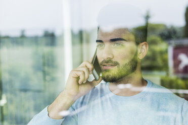 Junger Mann mit Mobiltelefon schaut aus dem Fenster - UUF007924