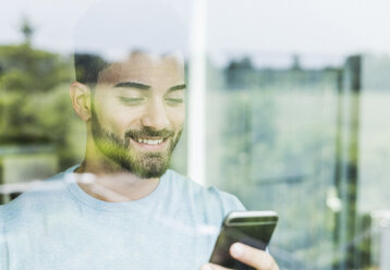 Smiling young man with cell phone behind windowpane - UUF007925