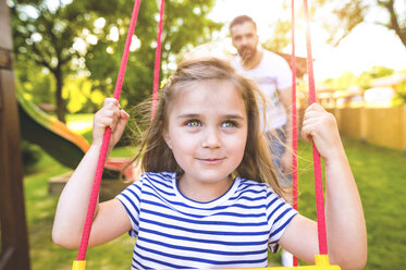 Porträt eines glücklichen kleinen Mädchens auf einer Schaukel mit Vater im Hintergrund - HAPF000557