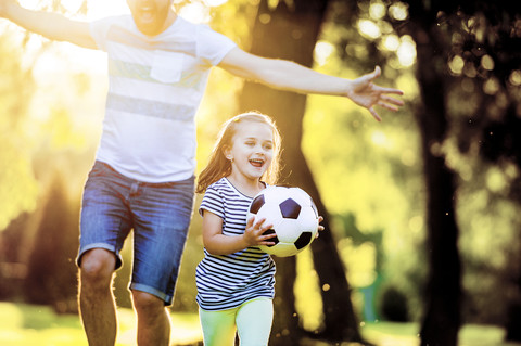 Glückliches kleines Mädchen mit Fußball, das mit seinem Vater in einem Park spielt, lizenzfreies Stockfoto