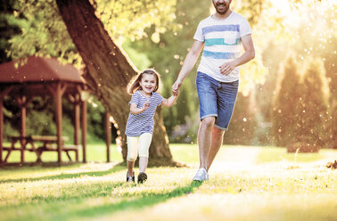 Portrait of smiling little girl running with her father hand in hand in a park - HAPF000577