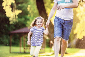 Porträt eines lächelnden kleinen Mädchens, das mit seinem Vater Hand in Hand in einem Park läuft - HAPF000578