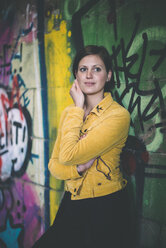 Porträt einer jungen Frau mit gelber Jacke, die vor einem Graffiti steht - DASF000057