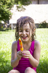 Porträt eines glücklichen kleinen Mädchens mit nassen Haaren, das mit einem Eislutscher auf einer Wiese sitzt - LVF005055