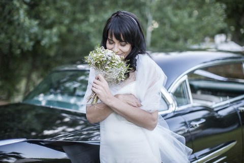 Die Braut riecht an ihrem Blumenstrauß vor einem Oldtimer, lizenzfreies Stockfoto