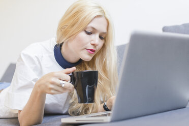 Blonde junge Frau liegt mit einer Tasse Kaffee auf der Couch und schaut auf ihren Laptop - FMOF000041