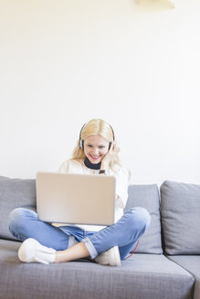 Blonde junge Frau sitzt mit Kopfhörern auf der Couch und schaut auf einen Laptop - FMOF000042