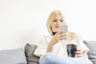 Blonde junge Frau sitzt mit einer Tasse Kaffee auf der Couch und schaut auf ihr Smartphone - FMOF000043