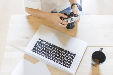 Frau sitzt mit Laptop und Kaffeetasse am Schreibtisch und hält eine Digitalkamera, Teilansicht - FMOF000046