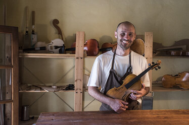 Lächelnder Geigenbauer mit einer Geige in seiner Werkstatt - ABZF000792
