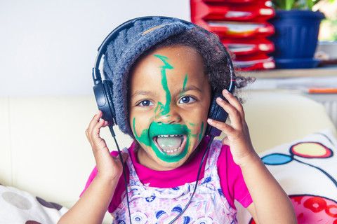 Porträt eines glücklichen kleinen Mädchens mit bemaltem Gesicht, das mit Kopfhörern Musik hört, lizenzfreies Stockfoto
