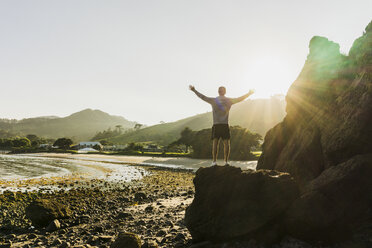 Neuseeland, Whangamata, Rückenansicht eines glücklichen Mannes, der auf einem Felsen steht und auf den Strand schaut - UUF007931