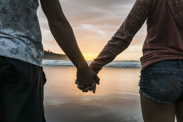 Couple standing hand in hand on the beach looking at sunset, partial view - UUF007961