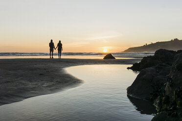 Silhouette eines Paares, das Hand in Hand am Strand steht und den Sonnenuntergang betrachtet - UUF007963