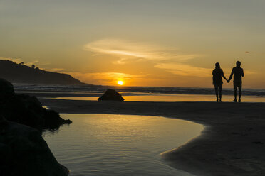 Silhouette eines Paares, das Hand in Hand am Strand steht und den Sonnenuntergang betrachtet - UUF007964