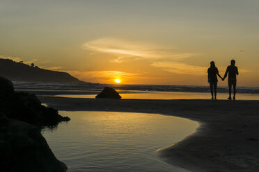 Silhouette of couple standing hand in hand on the beach looking at sunset - UUF007964