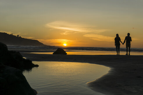Silhouette eines Paares, das Hand in Hand am Strand steht und den Sonnenuntergang betrachtet - UUF007964