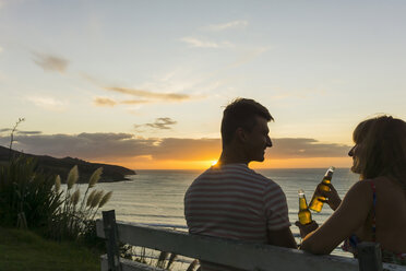 Couple sitting on bench at sunset toasting with beverages in glass bottles - UUF007967