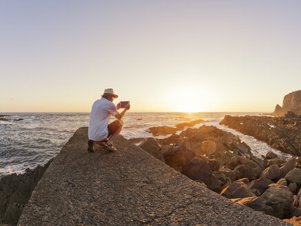 Portugal, älterer Mann, der bei Sonnenuntergang am Hafen auf einer Mauer sitzt und ein Foto mit einem digitalen Tablet macht - LAF001664