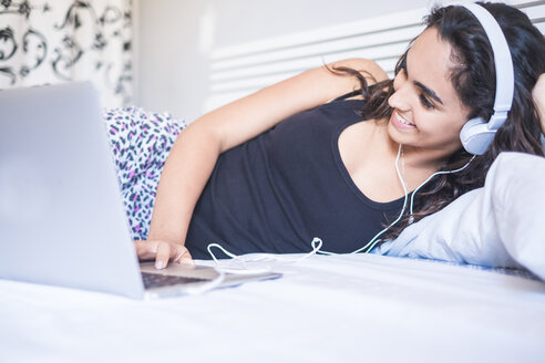 Teenager-Mädchen mit Kopfhörern auf dem Bett liegend mit Laptop - SIPF000599