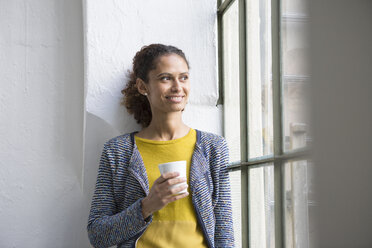 Junge Frau steht mit einer Tasse Kaffee am Fenster - RBF004663