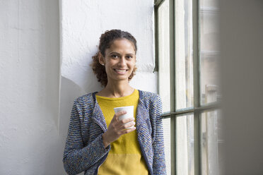 Junge Frau steht mit einer Tasse Kaffee am Fenster - RBF004664