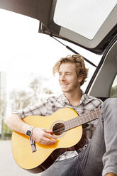 Porträt eines lächelnden jungen Mannes, der auf der Rückbank seines Autos sitzt und Gitarre spielt - PESF000182