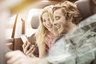 Lachendes junges Paar, das in einem Auto sitzt und gemeinsam auf ein Smartphone schaut - PESF000196