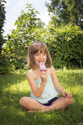 Portrait of happy little girl sitting on a meadow eating blueberry ice lolly - LVF005086