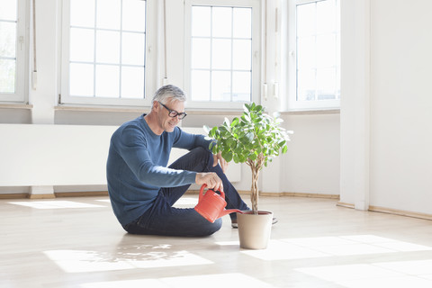 Mann sitzt auf dem Boden und gießt eine Pflanze in einer leeren Wohnung, lizenzfreies Stockfoto