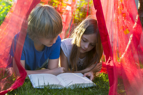 Schwester und Bruder liegen auf einer Wiese unter einem Moskitonetz und lesen gemeinsam ein Buch - SARF002812