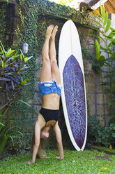 Young woman doing yoga, handstand in front of surfboard - KNTF000420