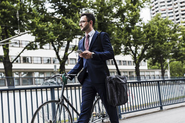 Businessman with bicycle walking in the city, using smart phone - UUF007994